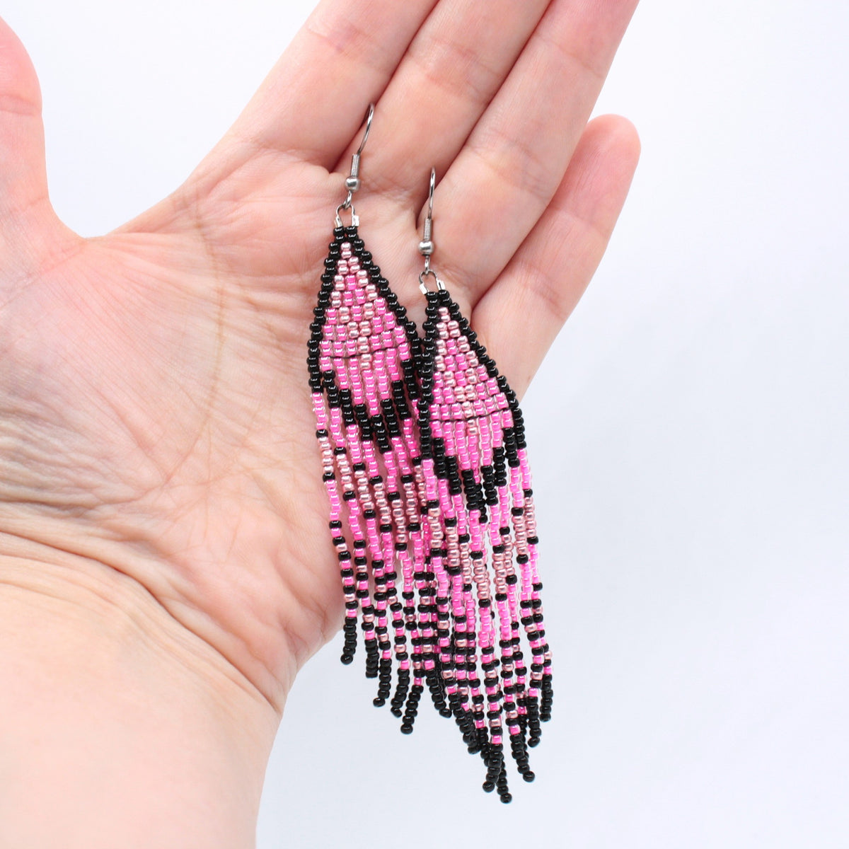 Hapsulliset pinkin sävyiset helmikorvakorut ovat esittelyssä roikkumassa kädessä. Seed bead fringe earrings in pink-toned are shown hanging from a hand.