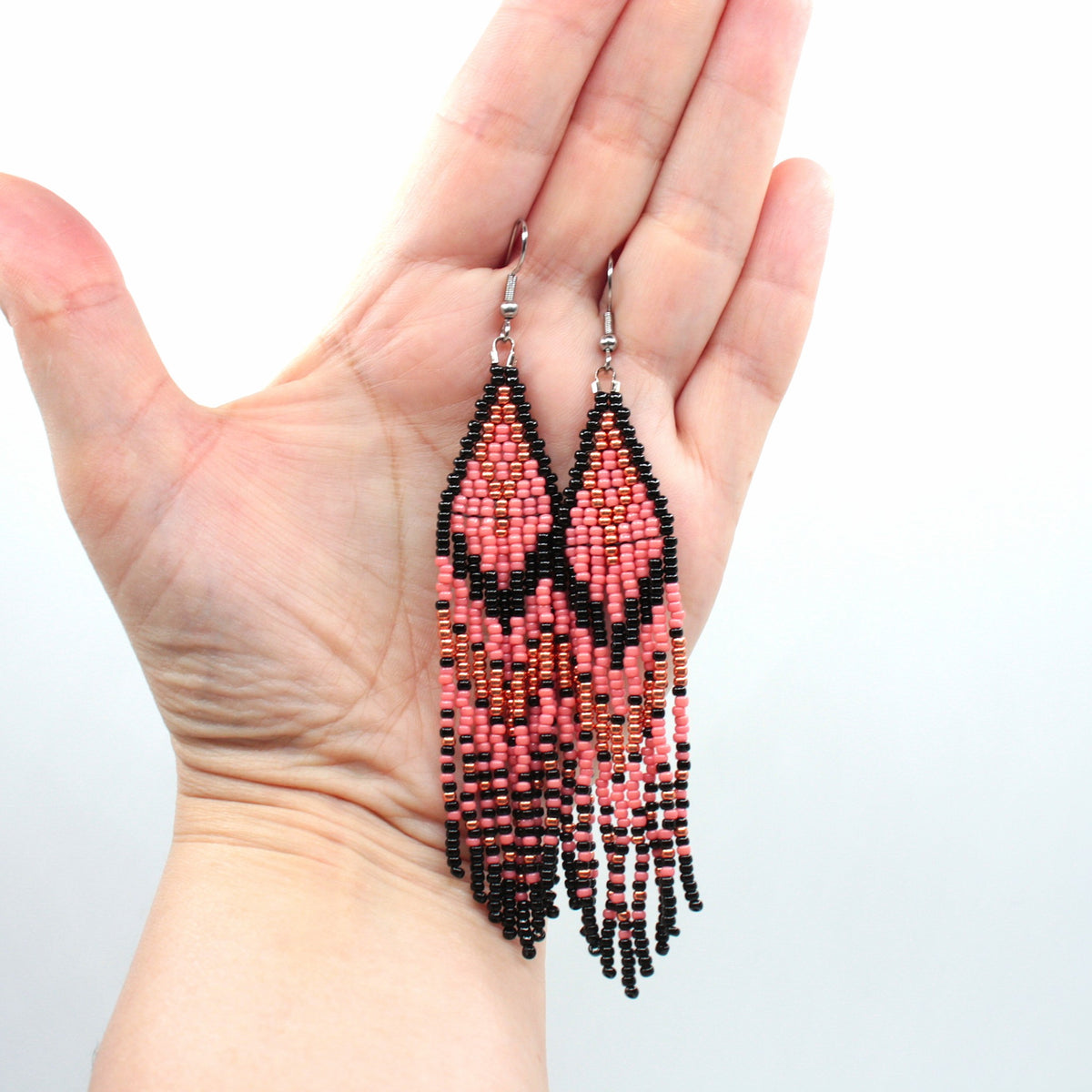 Vesimelonin pinkin ja mustan raidalliset hapsulliset lasihelmikorvakorut roikkuvat kädessä. Watermelon-pink and black striped seed bead fringe earrings hanging on the hand,