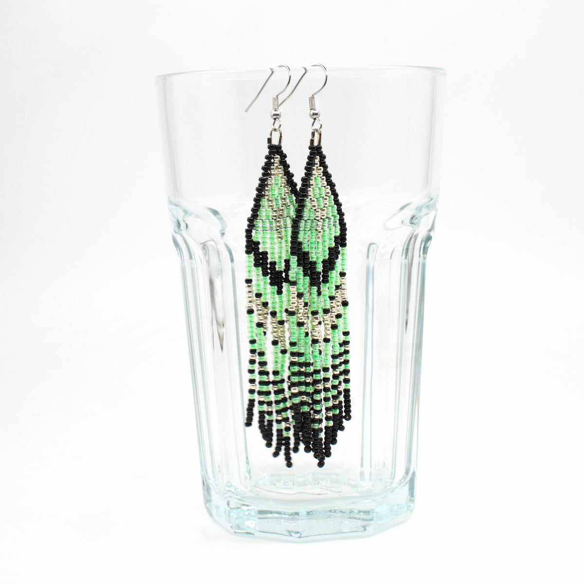 Raidalliset siemenhelmikorvakorut vihreän, musta ja hopean värisillä raidoilla. Tausta on valkoinen ja korvakorut roikkuvat juomalasin reunalla. Striped seed bead fringe earrings featuring green, black, and silver stripes. The background is white and the earrings are hanging from the rim of the drinking glass.