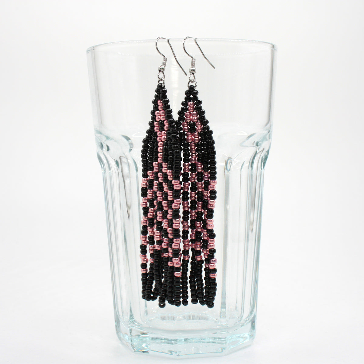 Hapsulliset lasihelmikorvakorut mustan ja pinkin väriset roikkuvat juomalasin reunalla. Black and pink seed bead fringe earrings hanging from the rim of the drinking class.