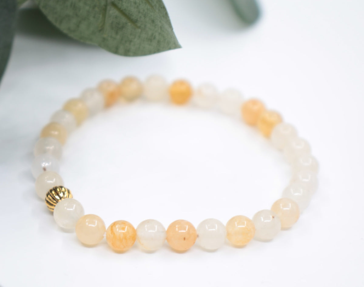 Keltainen jade joustava rannekoru kullan värisellä helmellä valkoisella alustalla. Taustalla vihreitä lehtiä. Yellow jade stretchy bracelet with gold color metal bead. Bracelet is on white background and there is green leafs.