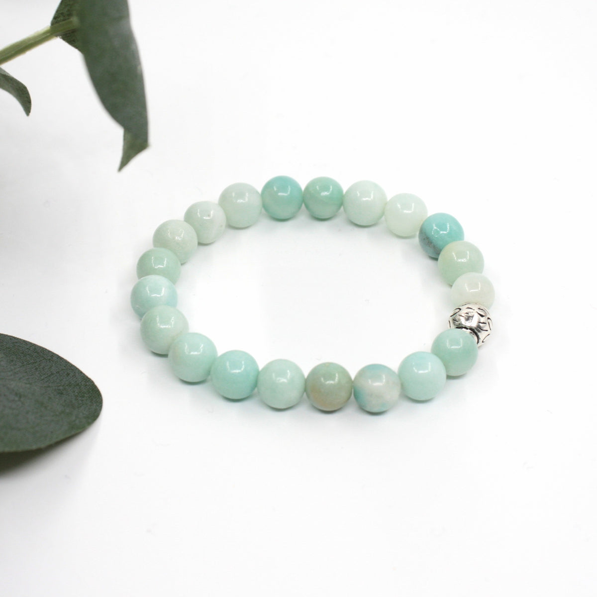 Amatsoniitti rannekoru valkoisella alustalla. Vasemmalla puolella kuvassa näkyy vähän vihreitä lehtiä. Amazonite bracelet on a white surface. A few green leaves are visible on the left side of the image.