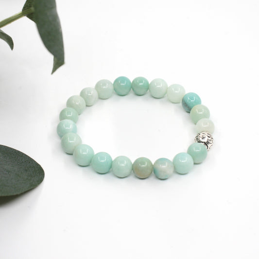 Amatsoniitti rannekoru valkoisella alustalla. Vasemmalla puolella kuvassa näkyy vähän vihreitä lehtiä. Amazonite bracelet on a white surface. A few green leaves are visible on the left side of the image.