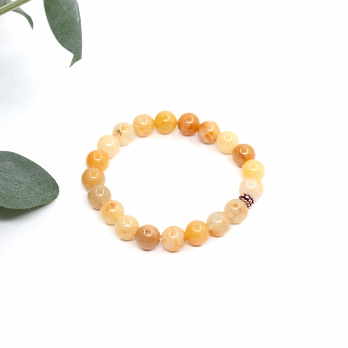 Keltainen jade rannekoru valkoisella alustalla. Vasemmalla puolella kuvassa näkyy vähän vihreitä lehtiä. Yellow jade bracelet on a white surface. A few green leaves are visible on the left side of the image.