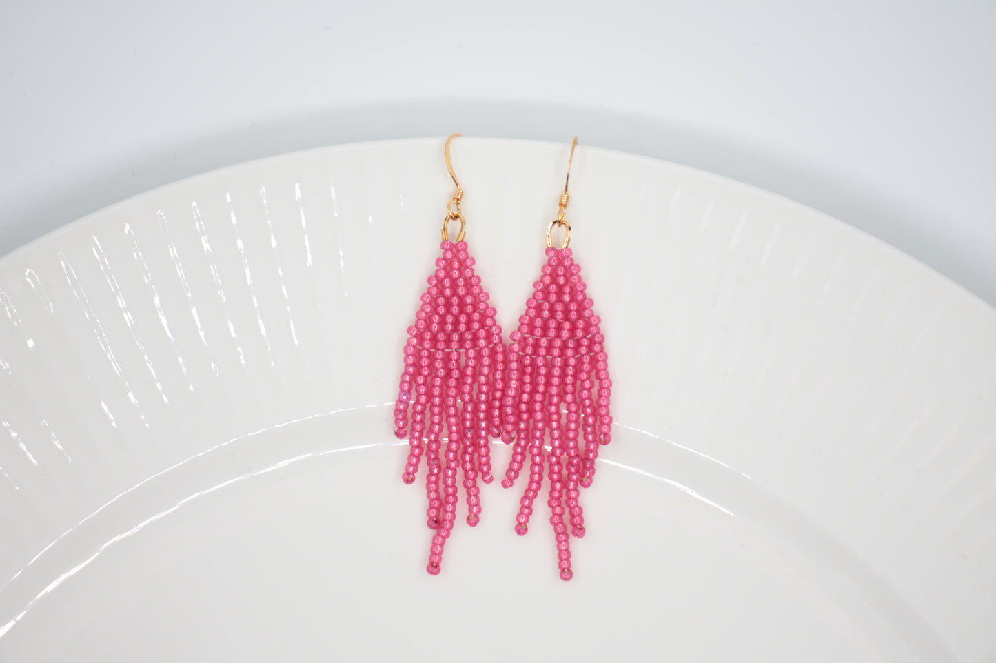 Roikkuvat hapsulliset helmikorvakorut värinä kimalteleva vadelma valkoisella taustalla. Seed bead fringe earrings sparkling raspberry- colored on the white background.