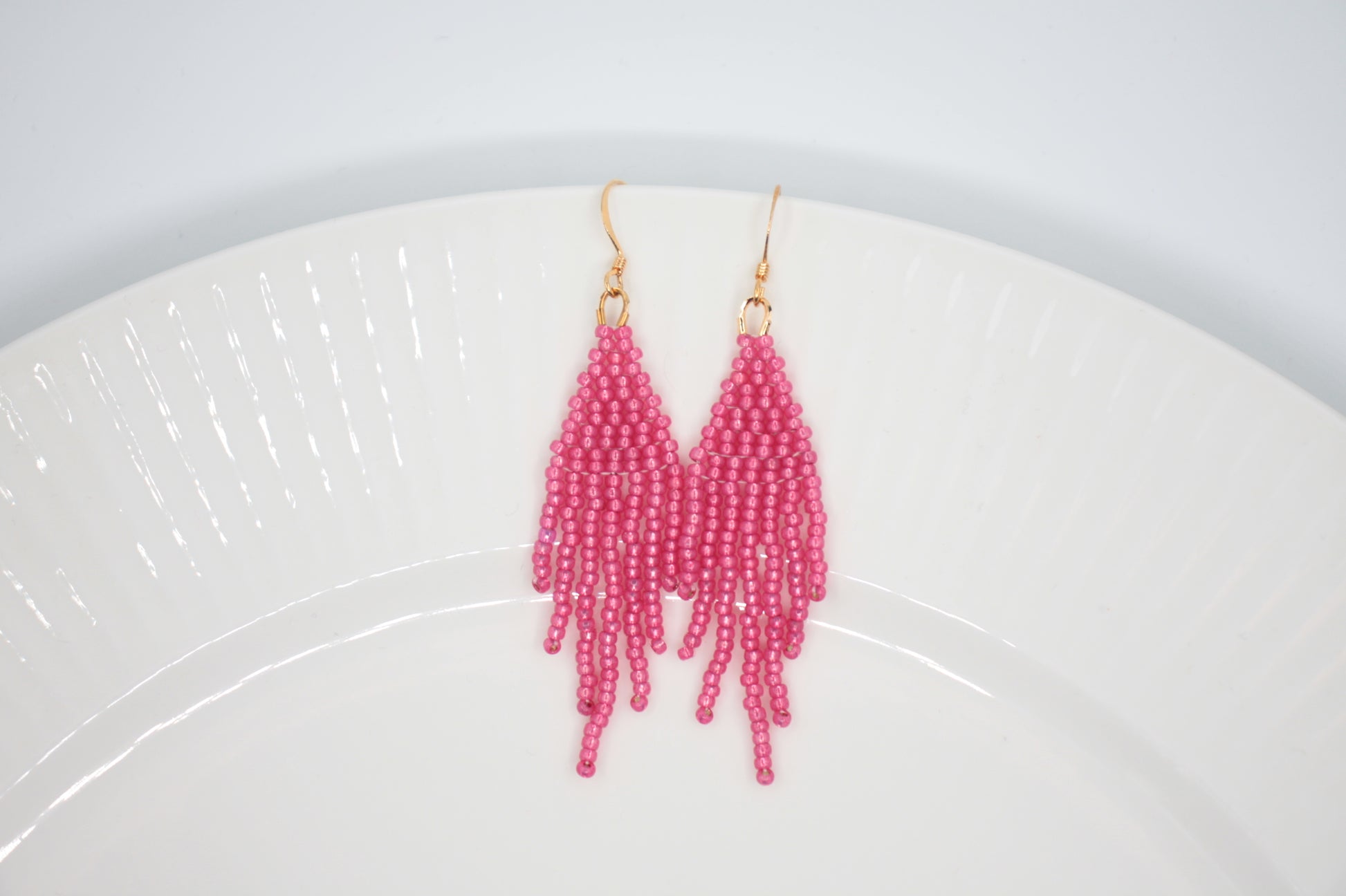 Roikkuvat hapsulliset helmikorvakorut värinä kimalteleva vadelma valkoisella taustalla. Seed bead fringe earrings sparkling raspberry- colored on the white background.