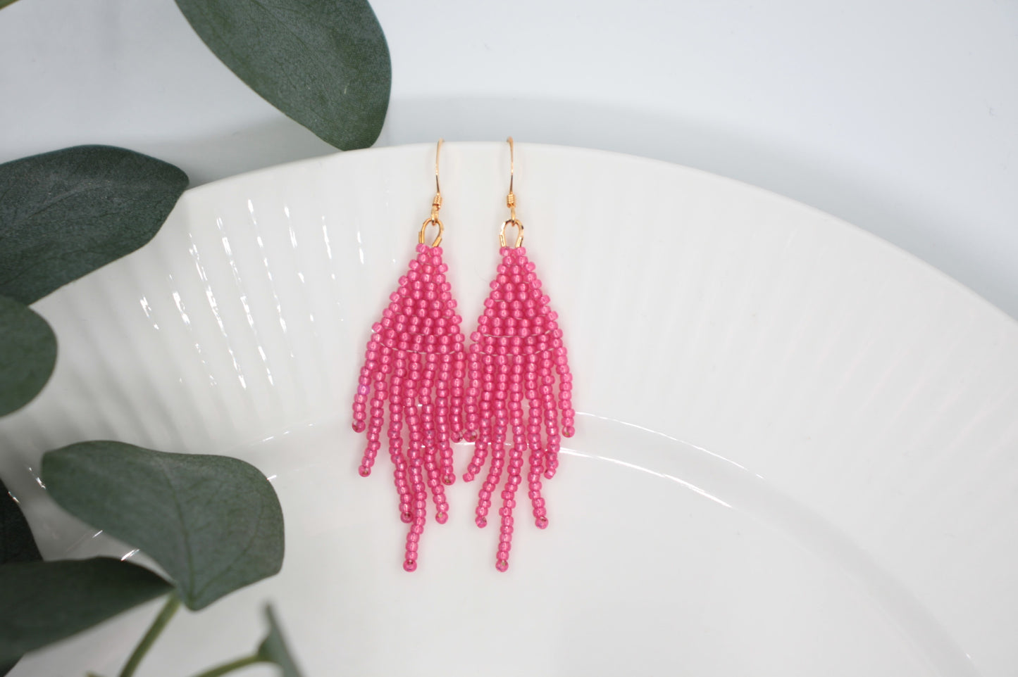 Roikkuvat hapsulliset helmikorvakorut värinä kimalteleva vadelma valkoisella taustalla. Seed bead fringe earrings sparkling raspberry- colored on the white background.