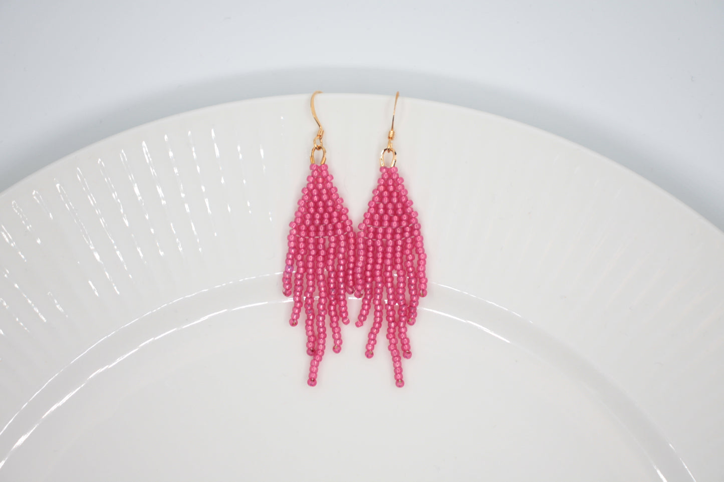 Roikkuvat hapsulliset helmikorvakorut värinä kimalteleva vadelma valkoisella taustalla. Seed bead fringe earrings sparkling raspberry- colored on the white background.