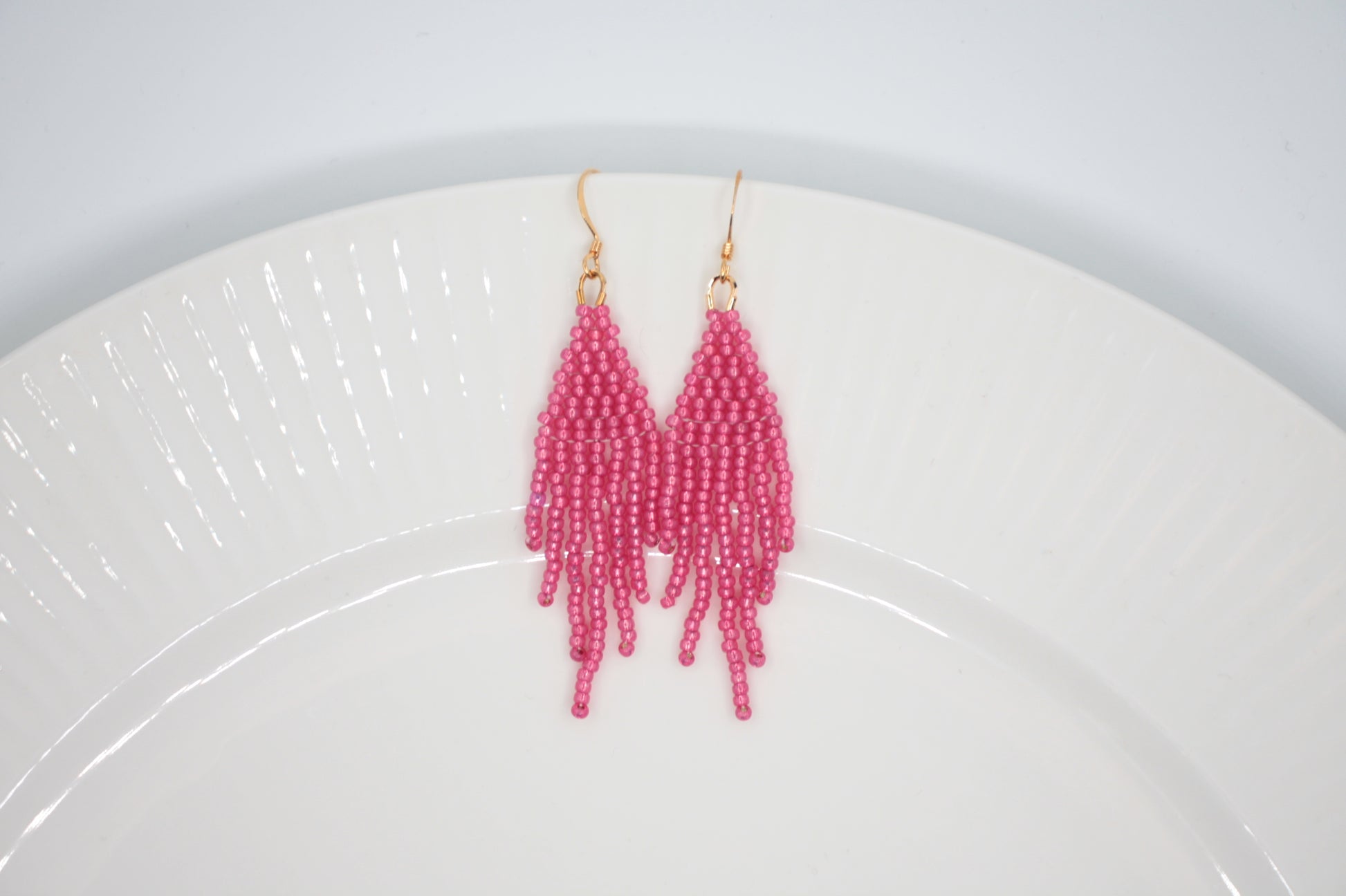 Roikkuvat hapsulliset helmikorvakorut värinä kimalteleva vadelma valkoisella taustalla. Seed bead fringe earrings sparkling raspberry- colored on the white background.