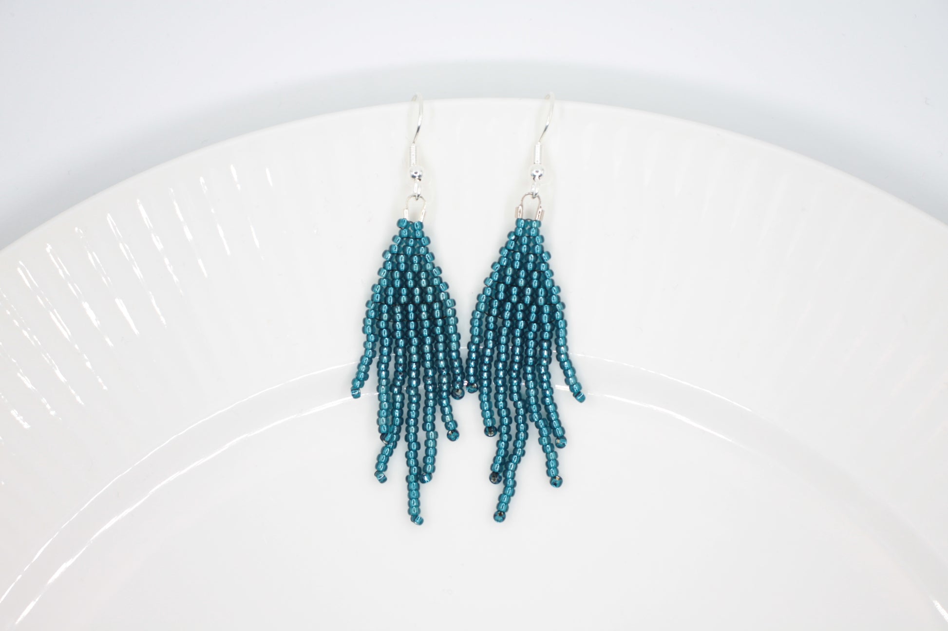 Sinisen safiirin väriset roikkuvat hapsulliset helmikorvakorut valkoisella alustalla. Seed bead fringe earrings blue sapphire-coloured on white background.