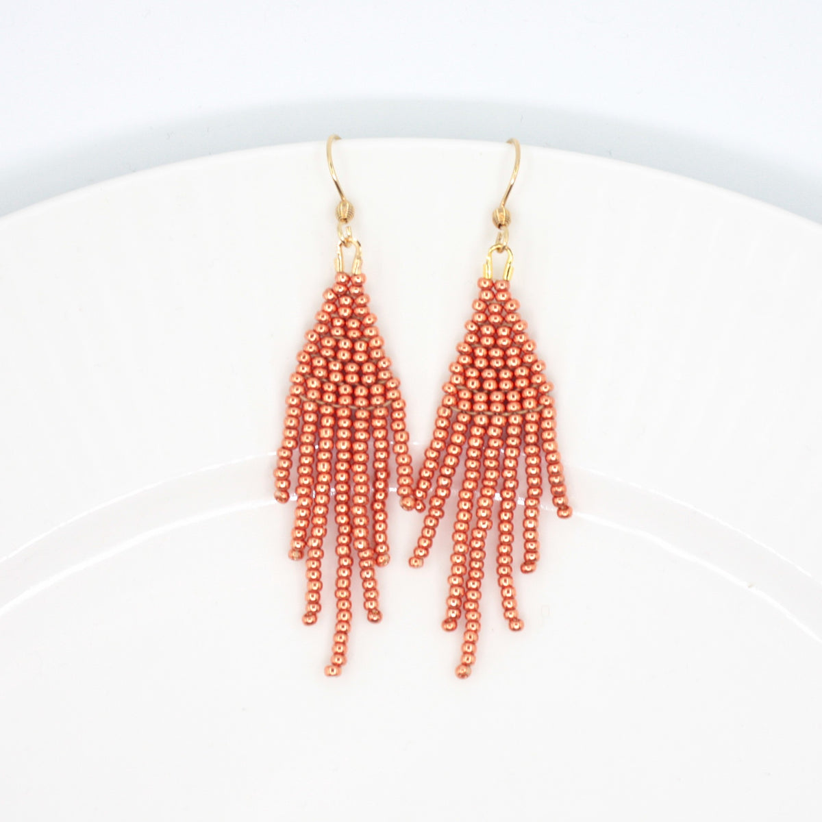 Hapsulliset helmikorvakorut värinä kupari valkoisella lautasella. Copper-colored beaded seed bead fringe earrings on a white plate. 