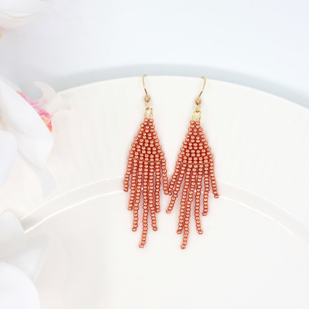 Hapsulliset helmikorvakorut värinä kupari valkoisella lautasella. Vasemmalla puolella näkyy valkoista kukkaa. Copper-colored beaded seed bead fringe earrings on a white plate. A white flower is visible on the left side.