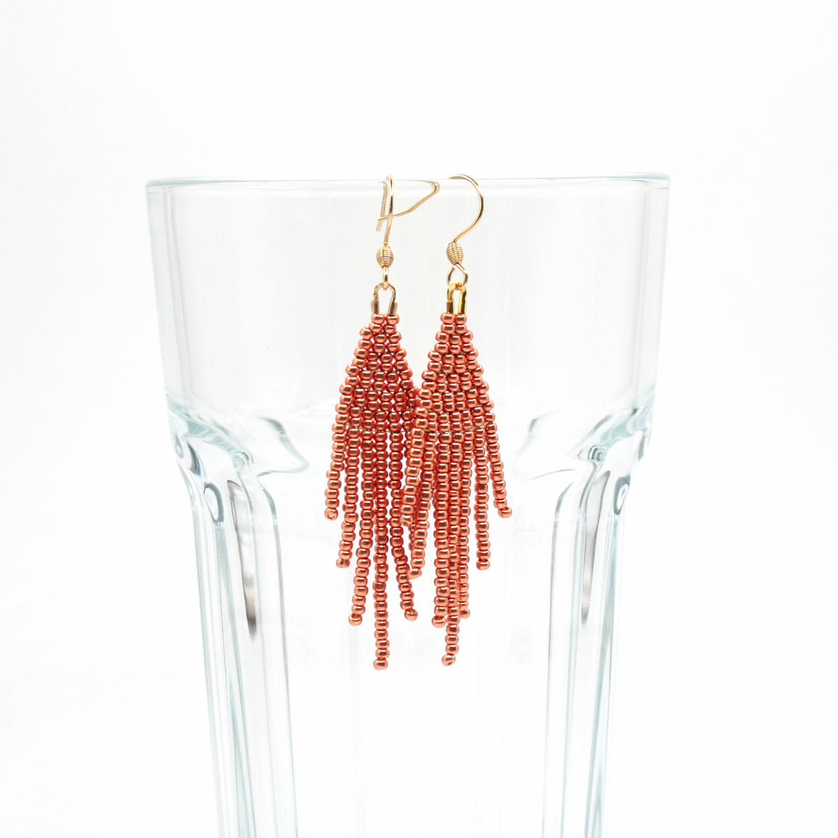 Hapsulliset helmikorvakorut värinä kupari roikkumassa juomalasin reunalla. Copper-colored beaded seed bead fringe earrings hanging from the rim of a drinking glass.