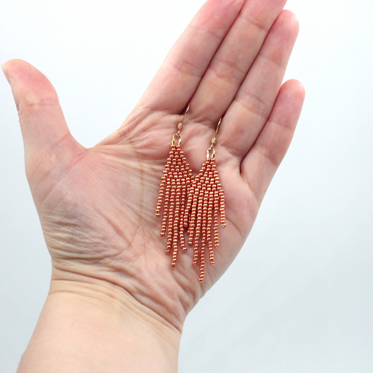 Hapsulliset helmikorvakorut värinä kupari roikkumassa kädessä. Copper-colored beaded seed bead fringe earrings hanging from the hand.