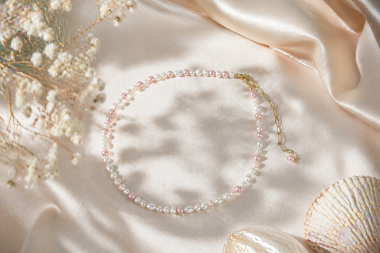 Handmade freshwater pearl necklace with pink and white glass beads and gold extension chain on soft neutral fabric background. Käsintehty makeanvedenhelmikaulakoru vaaleanpunaisilla ja valkoisilla lasihelmillä sekä kullanvärisellä jatkoketjulla vaalealla kangastaustalla.