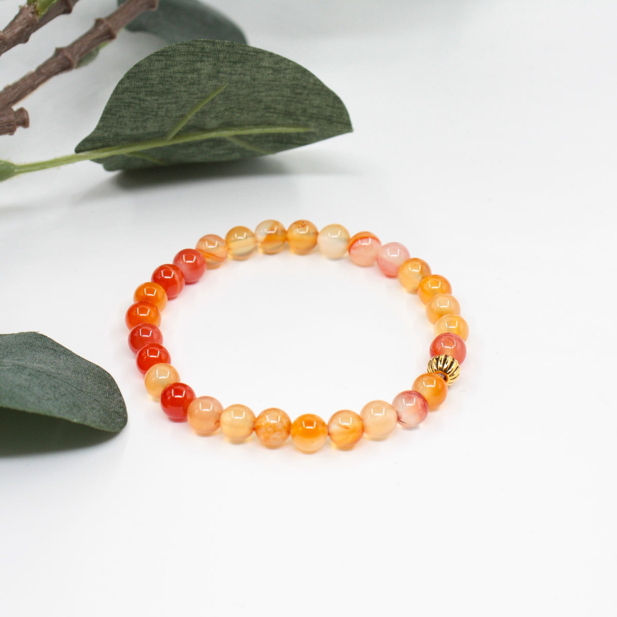 Karneoli rannekoru valkoisella alustalla. Taustalla vihreitä lehtiä. Carnelian bracelet on a white surface. Green leaves in the background.