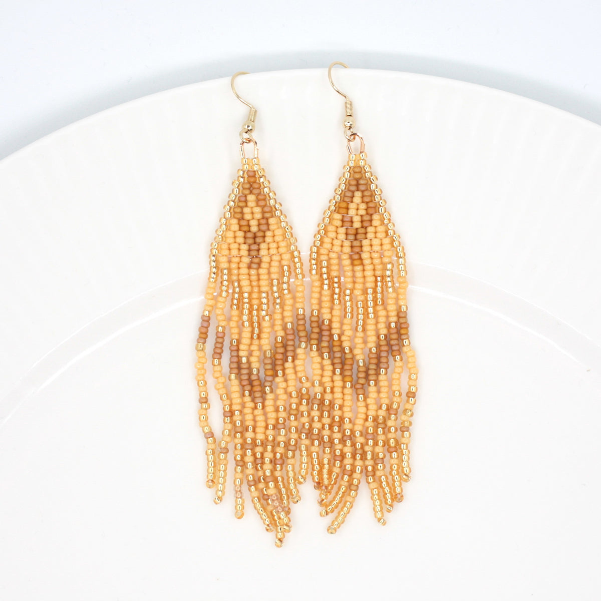 Hapsulliset beigen sävyiset helmikorvakorut valkoisen lautasen reunalla. Seed bead fringe earrings in beige toned resting on the edge of a white plate. 