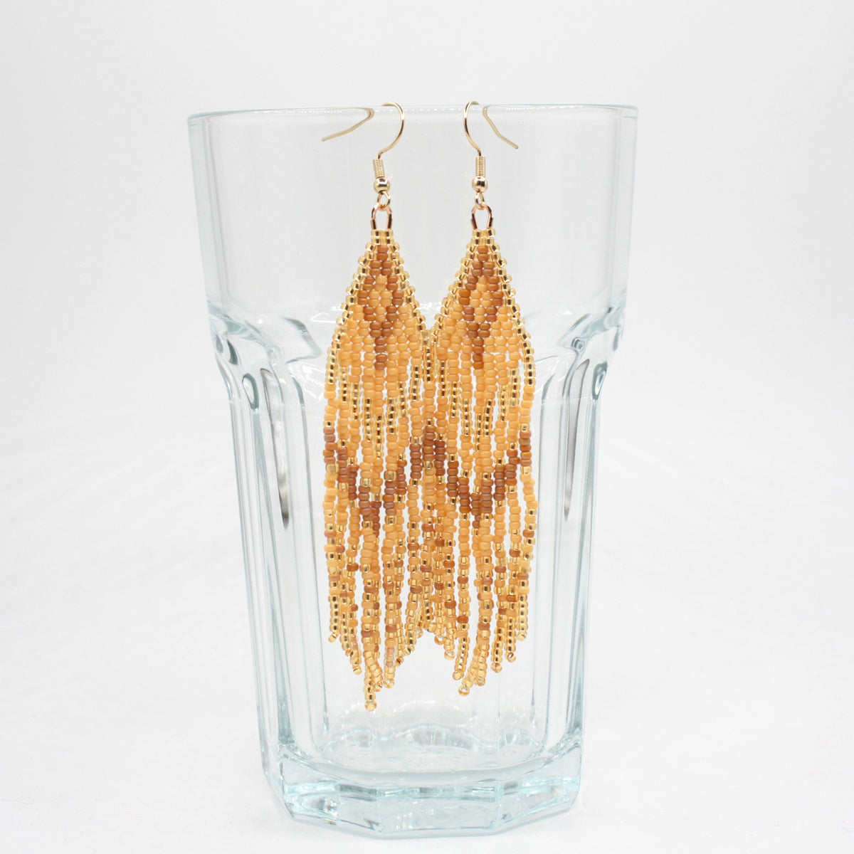 Hapsulliset beigen sävyiset helmikorvakorut roikkuvat juomalasin reunalla. Seed bead fringe earrings in beige toned on the rim of a drink glass.