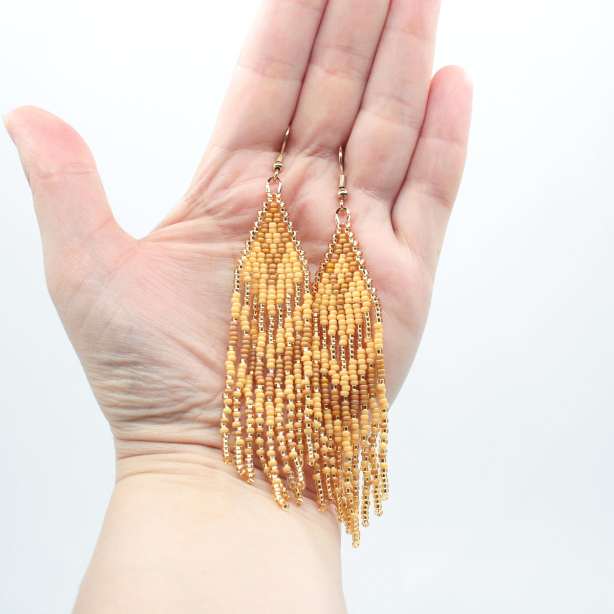 Hapsulliset beigen sävyiset helmikorvakorut ovat esittelyssä roikkumassa kädessä. Seed bead fringe earrings in beige-toned are shown hanging from a hand.