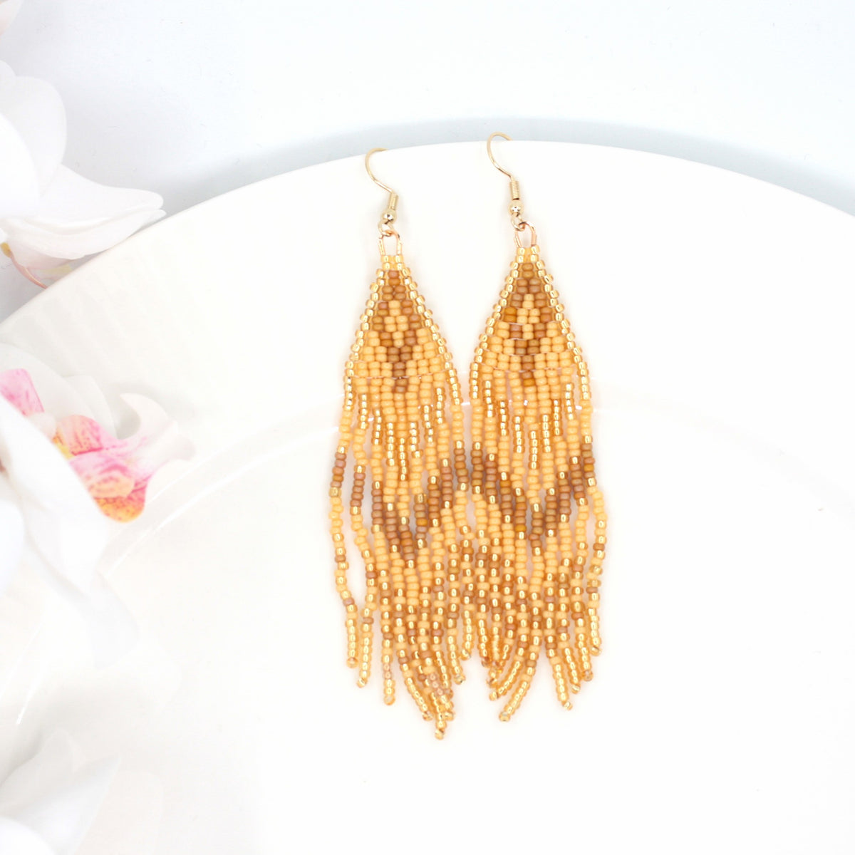 Hapsulliset beigen sävyiset helmikorvakorut valkoisen lautasen reunalla. Vasemmalla puolen näkyy valkoista kukkaa. Seed bead fringe earrings in beige toned resting on the edge of a white plate. A white flower is visible on the left side.