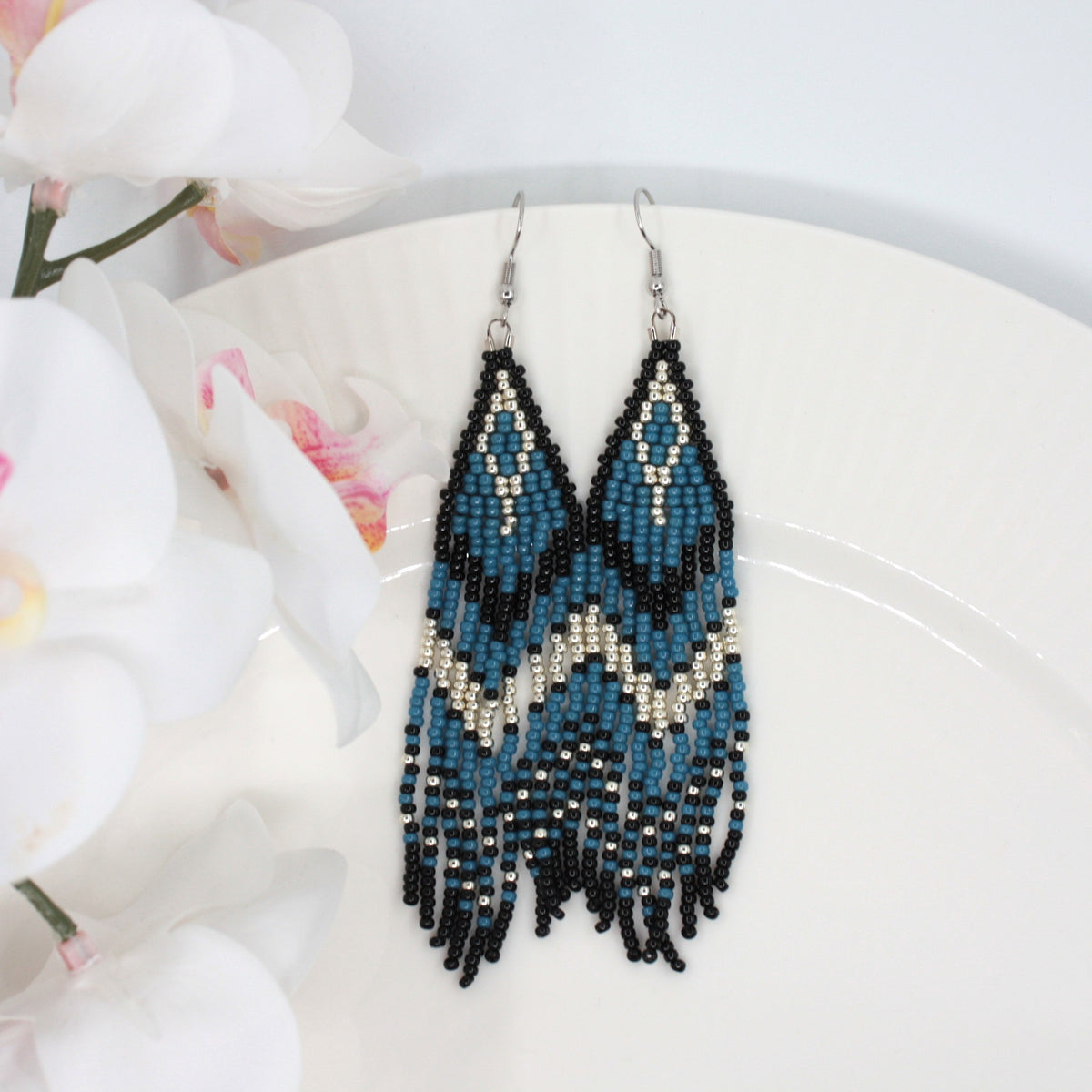 Hapsulliset katajan sinisen sävyiset helmikorvakorut valkoisen lautasen reunalla. Vasemmalla puolen näkyy valkoista kukkaa. Seed bead fringe earrings in juniper blue and black toned resting on the edge of a white plate. A white flower is visible on the left side.