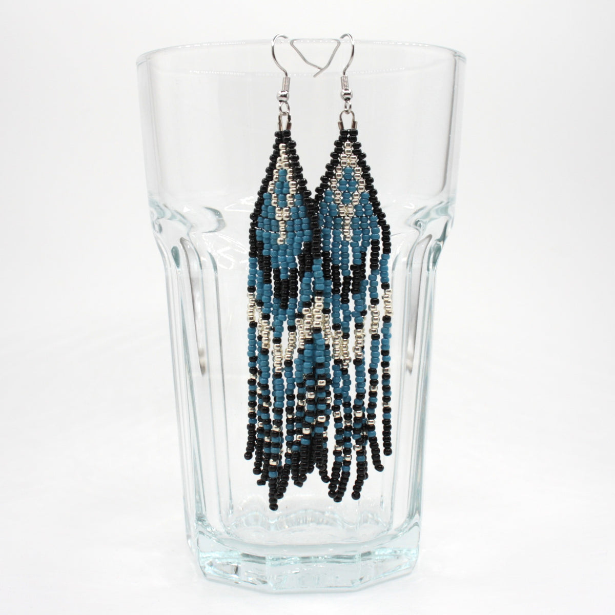 Hapsulliset katajan sinisen sävyiset helmikorvakorut roikkuvat juomalasin reunalla. Seed bead fringe earrings in juniper blue toned on the rim of a drink glass.