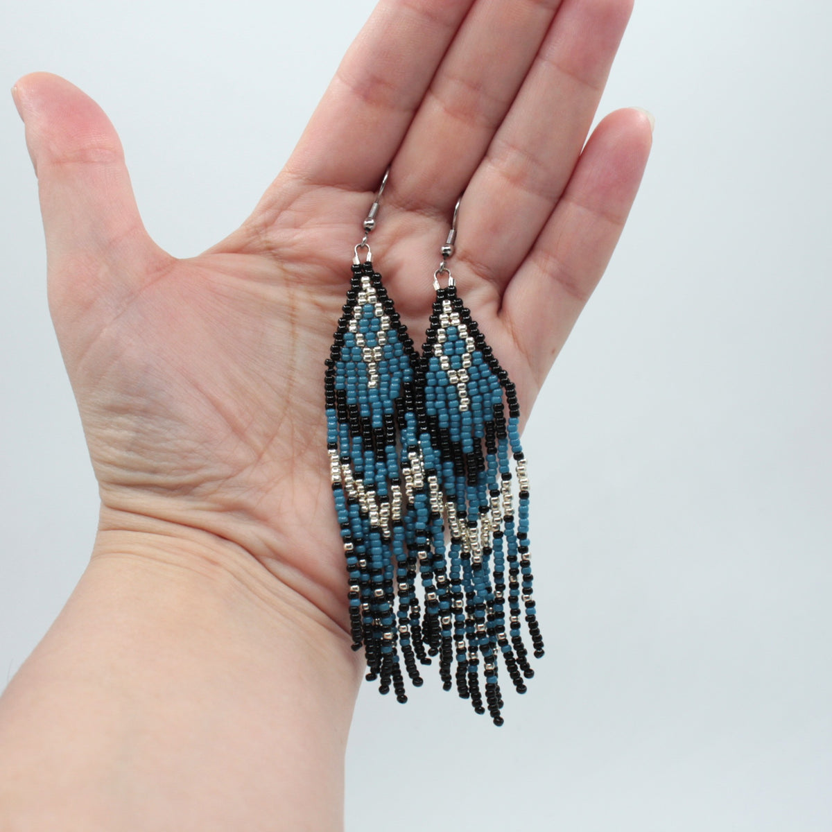Hapsulliset katajan sinisen sävyiset helmikorvakorut ovat esittelyssä roikkumassa kädessä. Seed bead fringe earrings in juniper blue-toned are shown hanging from a hand.