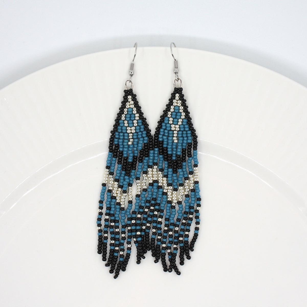 Hapsulliset katajan sinisen sävyiset helmikorvakorut valkoisen lautasen reunalla. Seed bead fringe earrings in juniper blue and black toned resting on the edge of a white plate. 