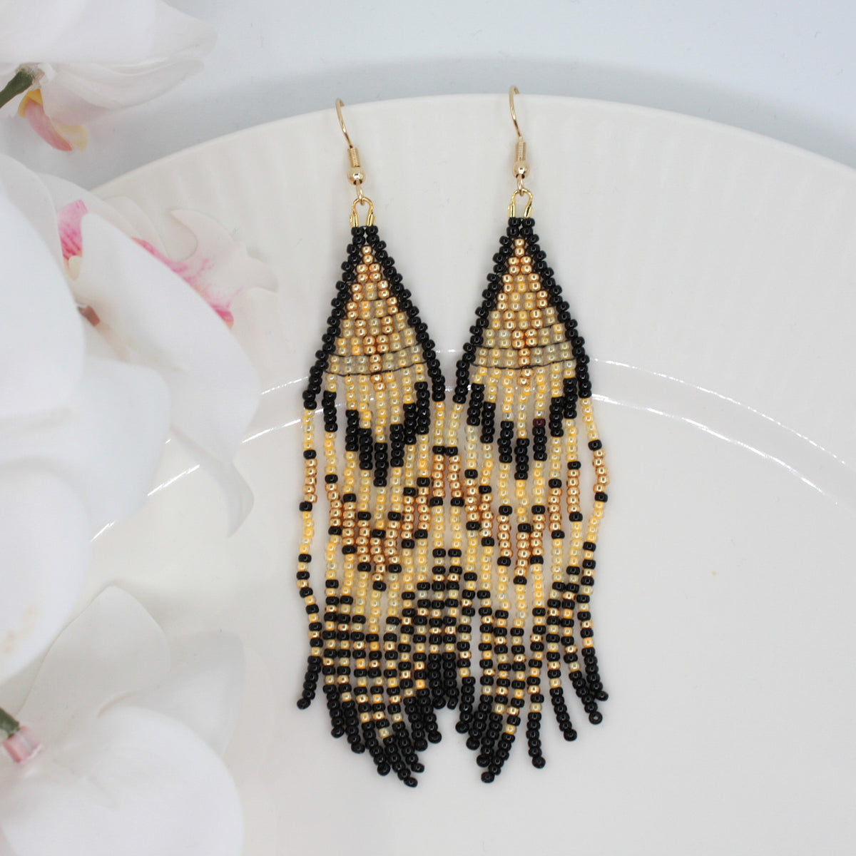 Hapsulliset keltaisen sävyiset helmikorvakorut valkoisen lautasen reunalla. Vasemmalla puolen näkyy valkoista kukkaa. Seed bead fringe earrings in yellow and black toned resting on the edge of a white plate. A white flower is visible on the left side.