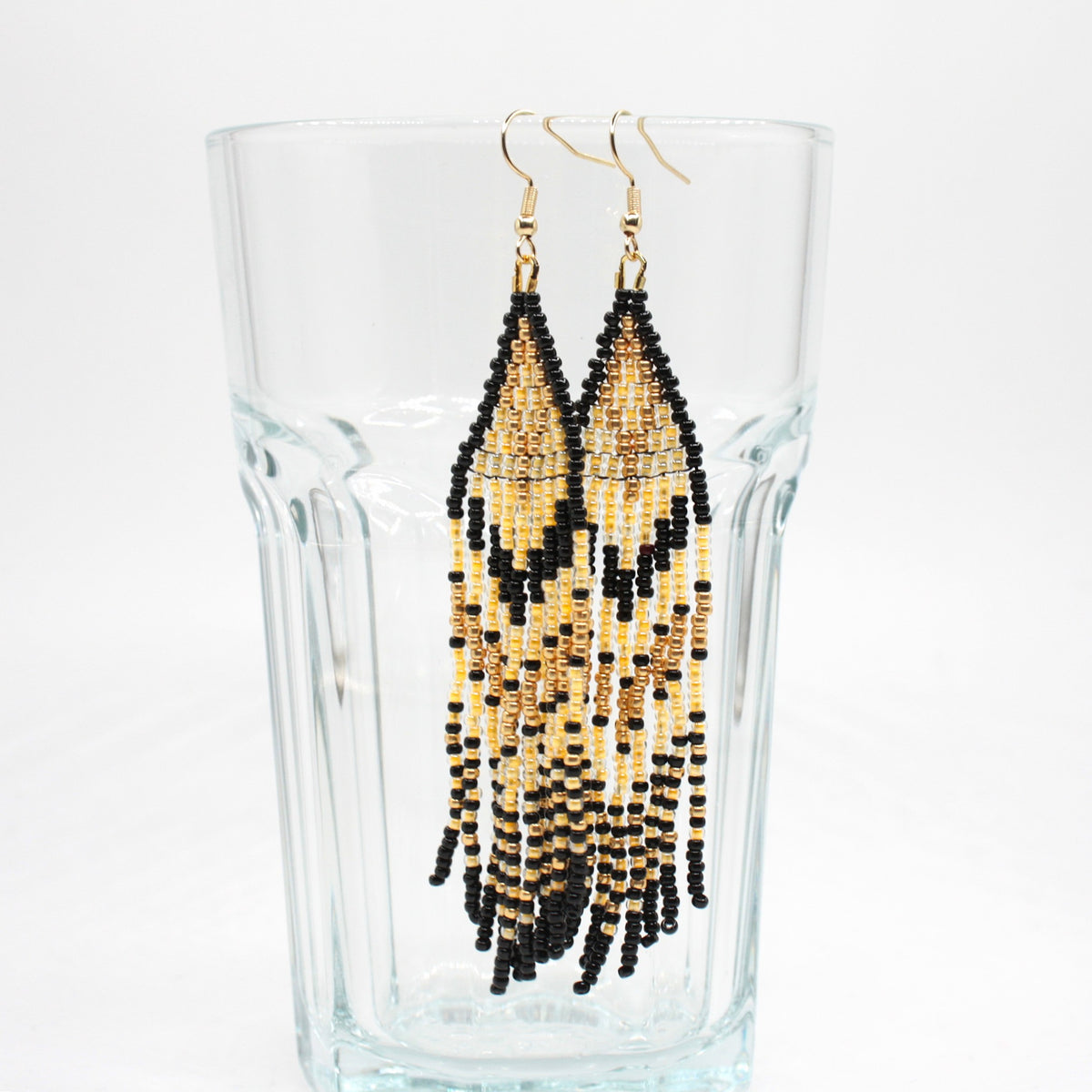 Hapsulliset keltaisen sävyiset helmikorvakorut roikkuvat juomalasin reunalla. Seed bead fringe earrings in yellow toned on the rim of a drink glass.