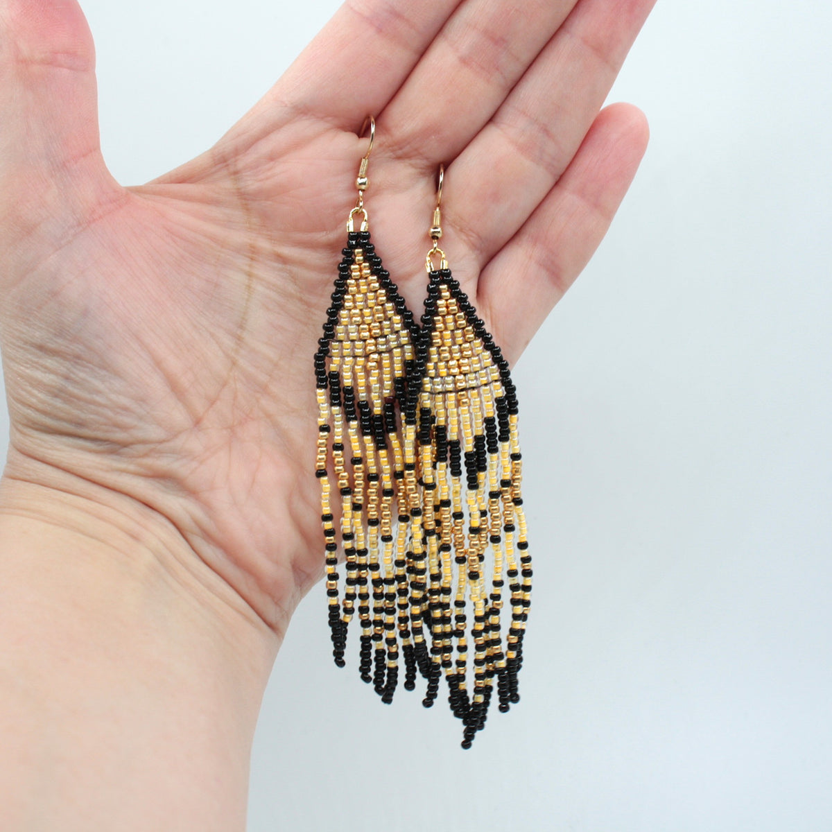 Hapsulliset keltaisen sävyiset helmikorvakorut ovat esittelyssä roikkumassa kädessä. Seed bead fringe earrings in yellow-toned are shown hanging from a hand.