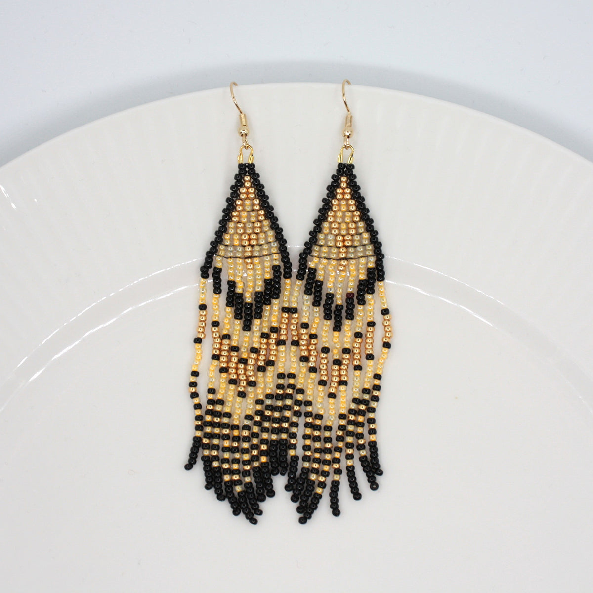 Hapsulliset keltaisen sävyiset helmikorvakorut valkoisen lautasen reunalla. Seed bead fringe earrings in yellow and black toned resting on the edge of a white plate. 
