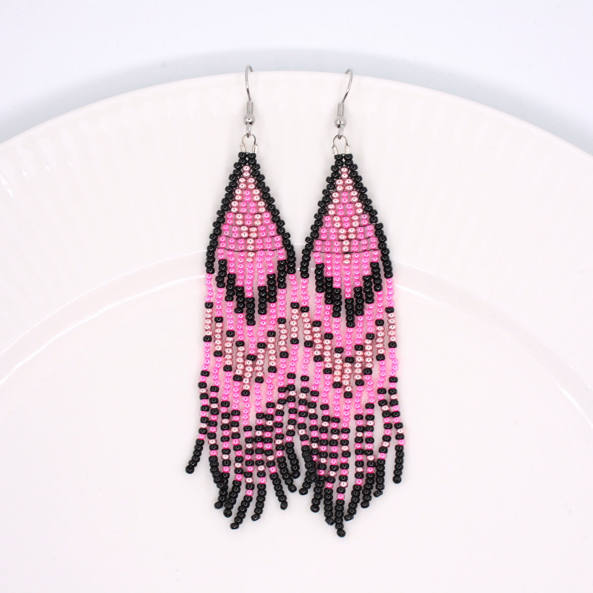 Hapsulliset pinkin sävyiset helmikorvakorut valkoisen lautasen reunalla. Seed bead fringe earrings in pink toned resting on the edge of a white plate. 
