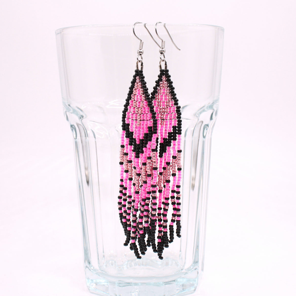 Hapsulliset pinkin sävyiset helmikorvakorut roikkuvat juomalasin reunalla. Seed bead fringe earrings in pink toned on the rim of a drink glass.