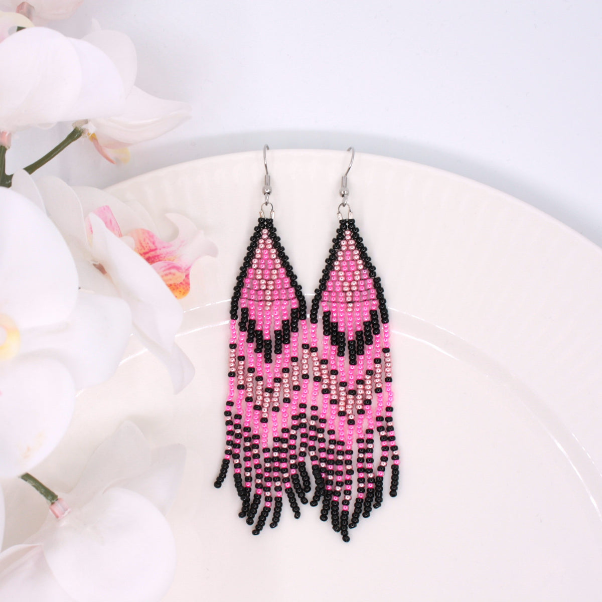 Hapsulliset pinkin sävyiset helmikorvakorut valkoisen lautasen reunalla. Vasemmalla puolen näkyy valkoista kukkaa. Seed bead fringe earrings in pink toned resting on the edge of a white plate. A white flower is visible on the left side.