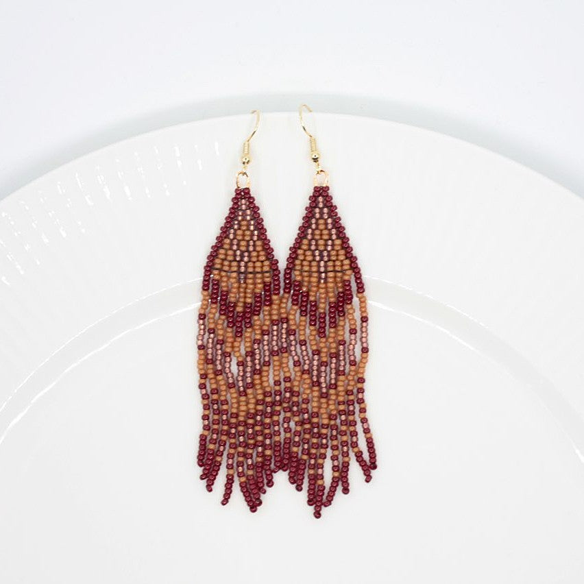 Hapsulliset ruskean sävyiset helmikorvakorut valkoisen lautasen reunalla. Seed bead fringe earrings in brown toned resting on the edge of a white plate. 
