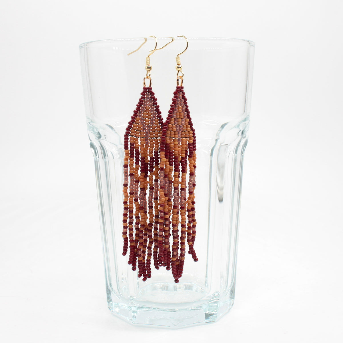 Hapsulliset ruskean sävyiset helmikorvakorut roikkuvat juomalasin reunalla. Seed bead fringe earrings in brown toned  on the rim of a drink glass. 