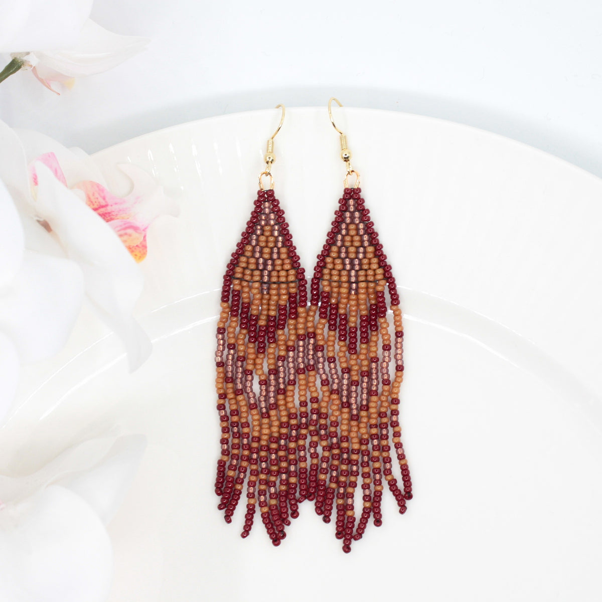 Hapsulliset ruskean sävyiset helmikorvakorut valkoisen lautasen reunalla. Vasemmalla puolen näkyy valkoista kukkaa. Seed bead fringe earrings in brown toned resting on the edge of a white plate. A white flower is visible on the left side.