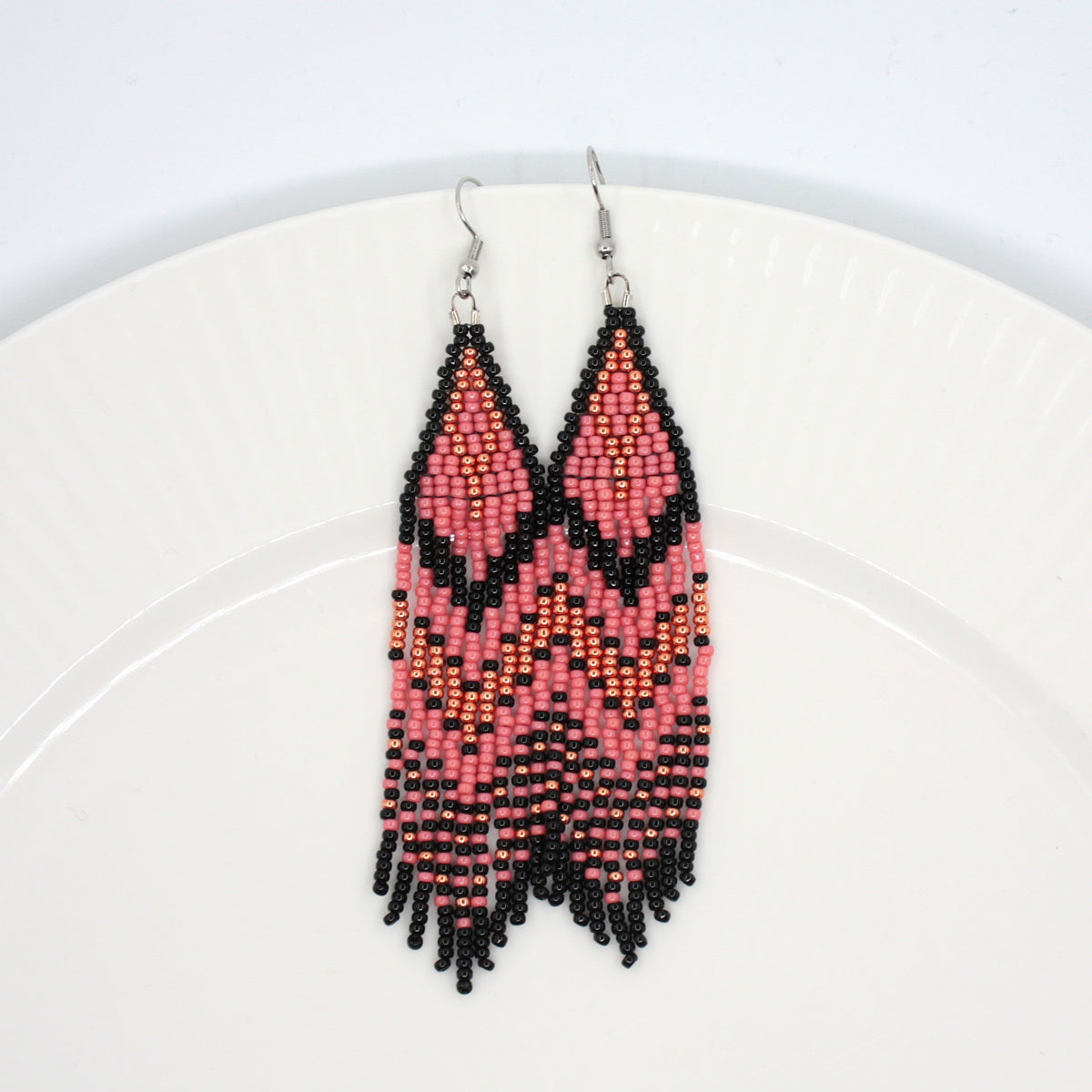 Vesimelonin pinkin ja mustan raidalliset hapsulliset lasihelmikorvakorut valkoisella lautasella. Watermelon-pink and black striped seed bead fringe earrings on a white plate. 