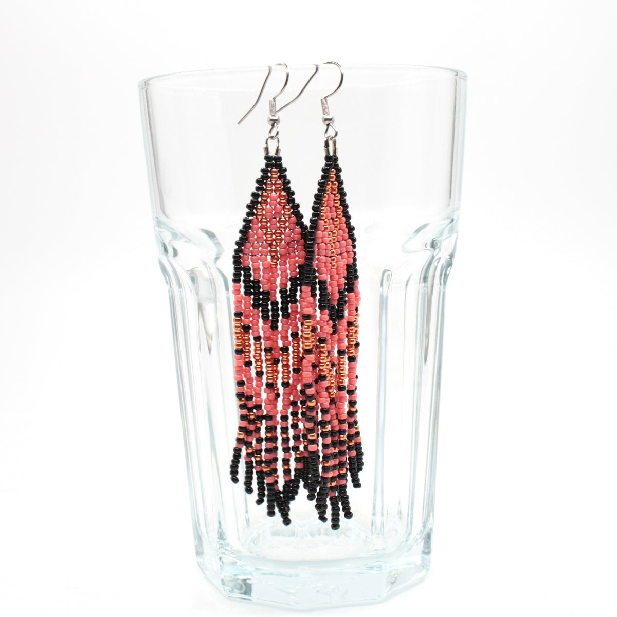 Vesimelonin pinkin ja mustan raidalliset hapsulliset lasihelmikorvakorut roikkuvat juomalasin reunalla. Watermelon-pink and black striped seed bead fringe earrings hanging from the rim of the drinking glass.