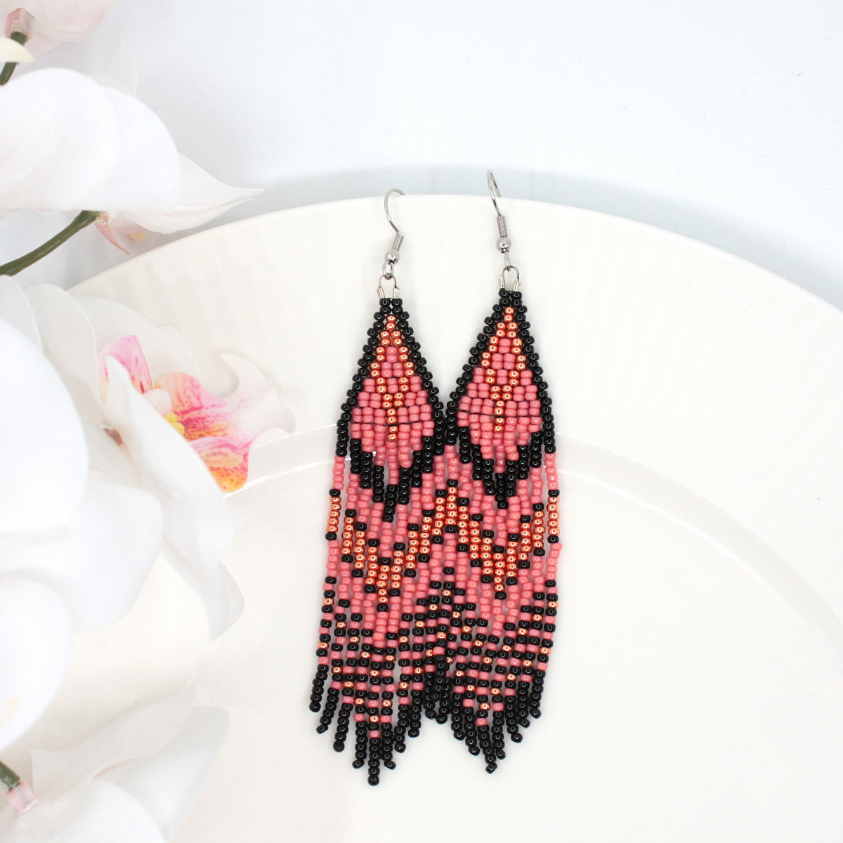 Vesimelonin pinkin ja mustan raidalliset hapsulliset lasihelmikorvakorut valkoisella lautasella. Vasemmalla puolella on valkoisia kukkia. Watermelon-pink and black striped seed bead fringe earrings on a white plate. There are white flowers on the left side.
