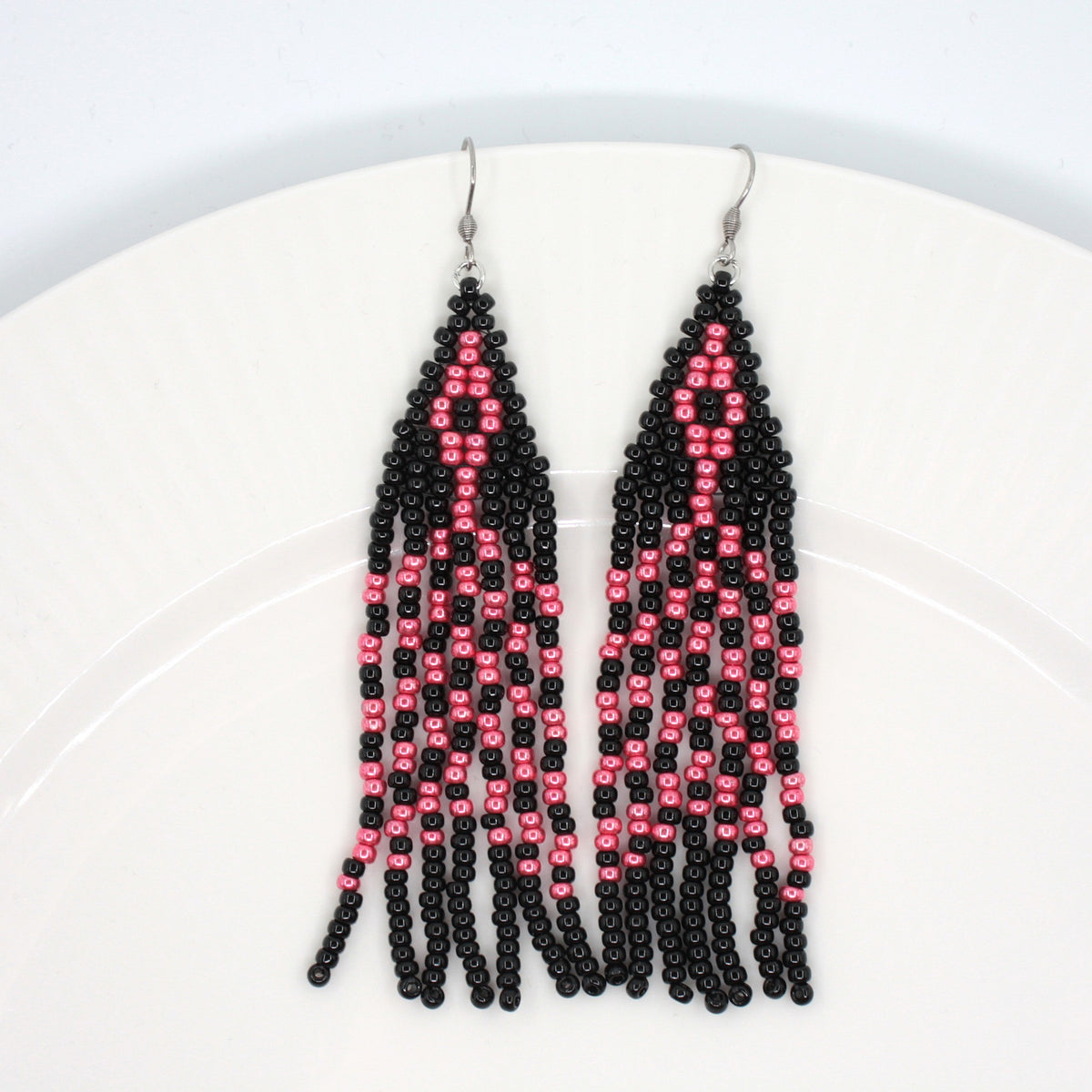 Hapsulliset lasihelmikorvakorut mustan ja  vadelman väriset valkoisella lautasella. Black and raspberry seed bead fringe earrings on the white plate.