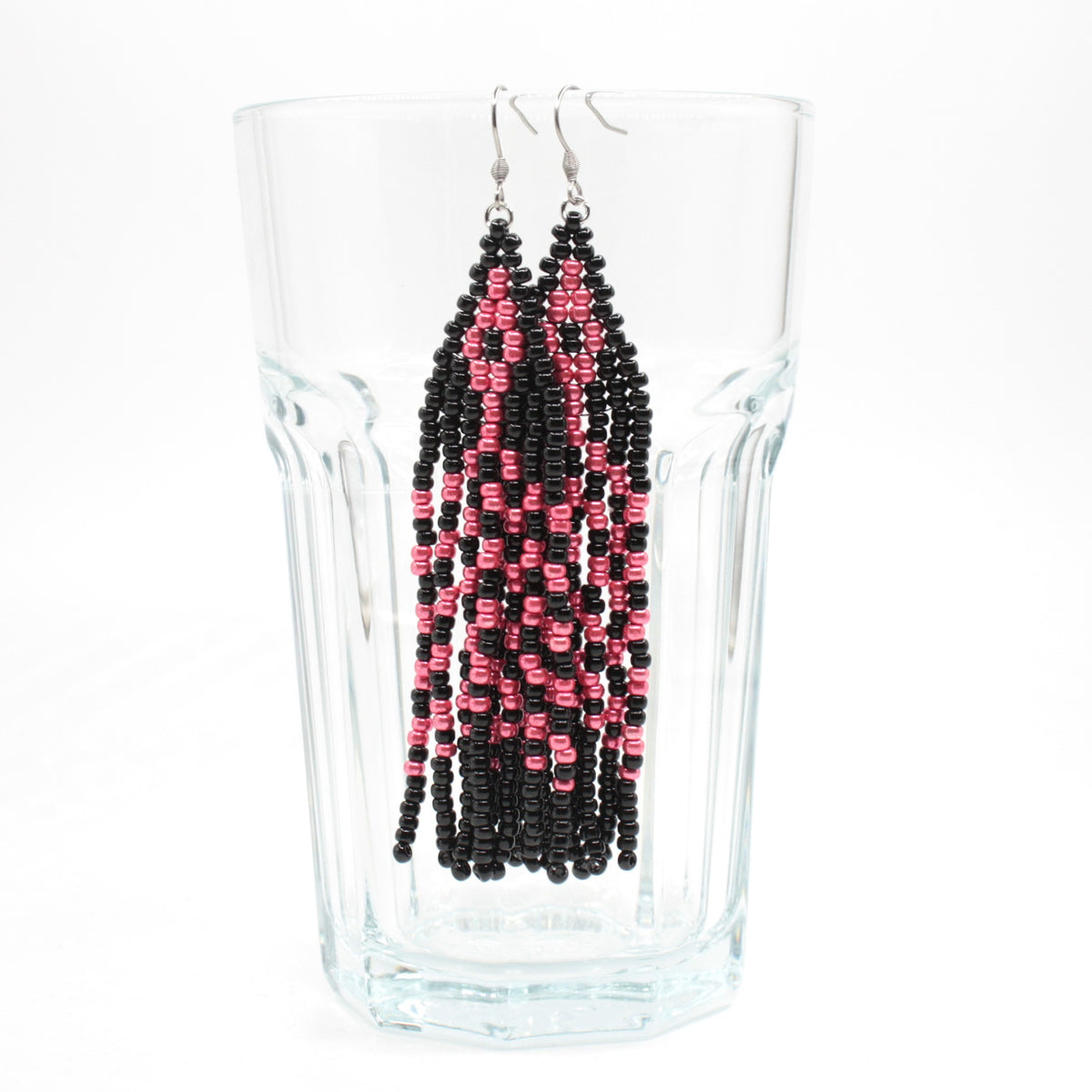 Hapsulliset lasihelmikorvakorut mustan ja vadelman väriset roikkuvat juomalasin reunalla. Black and raspberry seed bead fringe earrings hanging from the rim of the drinking class.