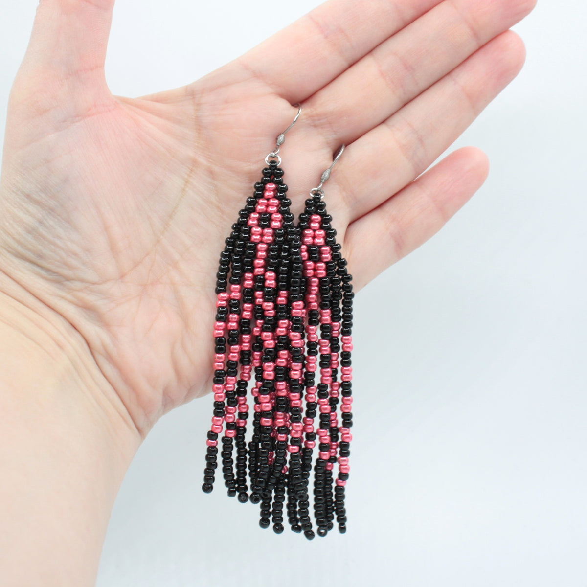 Hapsulliset lasihelmikorvakorut mustan ja vadelman väriset roikkuvat esillä kädessä. Black and raspberry seed bead fringe earrings hanging from the hand.