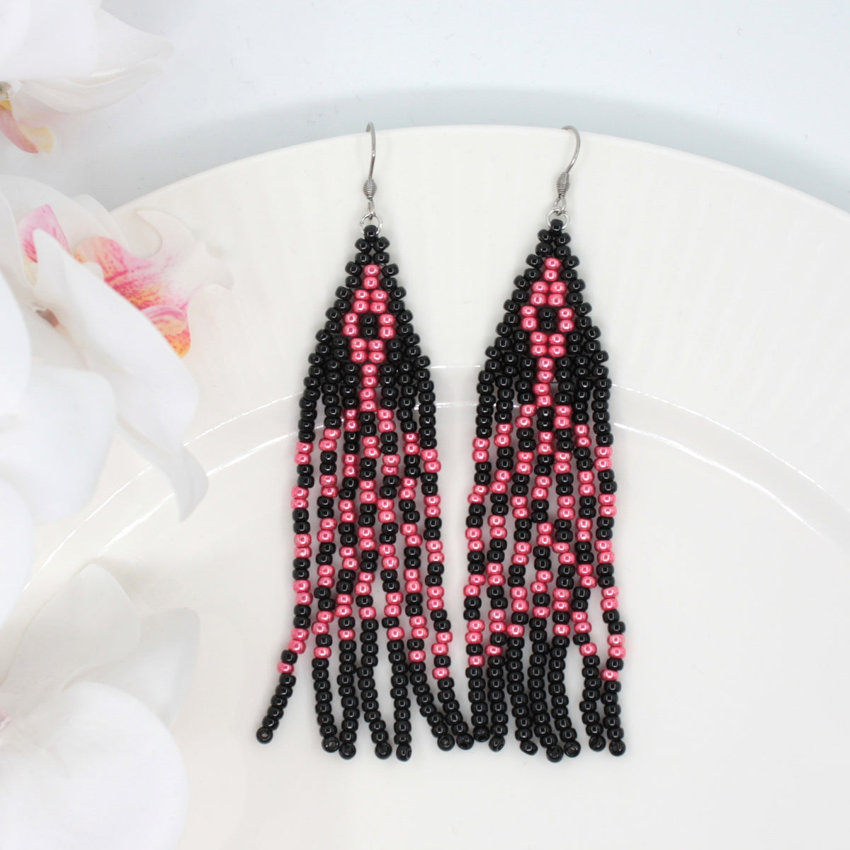 Hapsulliset lasihelmikorvakorut mustan ja  vadelman väriset valkoisella alustalla. Vasemmalla puolen valkoisia kukkia, jossa vähän myös pinkkiä. Black and raspberry seed bead fringe earrings on white surface. Left side is white flowers with little bit pink.