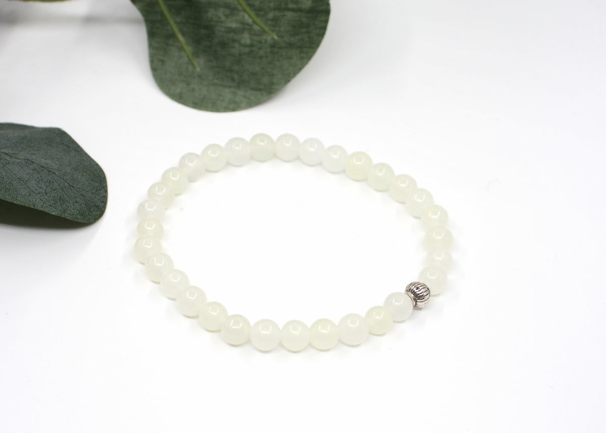 Jade rannekoru hopean värisellä helmellä. Alusta on valkoinen ja taustalla näkyy vihreitä lehtiä. Jade bracelet with one silver color bead. Bracelet is on white setting and background is green leaves.
