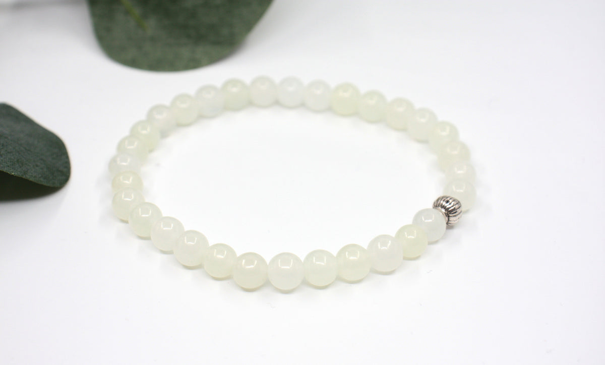 Jade rannekoru hopean värisellä helmellä. Alusta on valkoinen ja taustalla näkyy vihreitä lehtiä. Jade bracelet with one silver color bead. Bracelet is on white setting and background is green leaves.