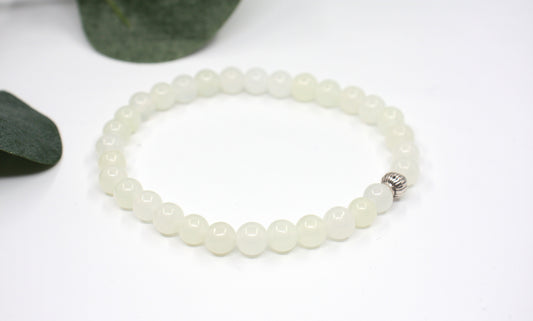Jade rannekoru hopean värisellä helmellä. Alusta on valkoinen ja taustalla näkyy vihreitä lehtiä. Jade bracelet with one silver color bead. Bracelet is on white setting and background is green leaves.