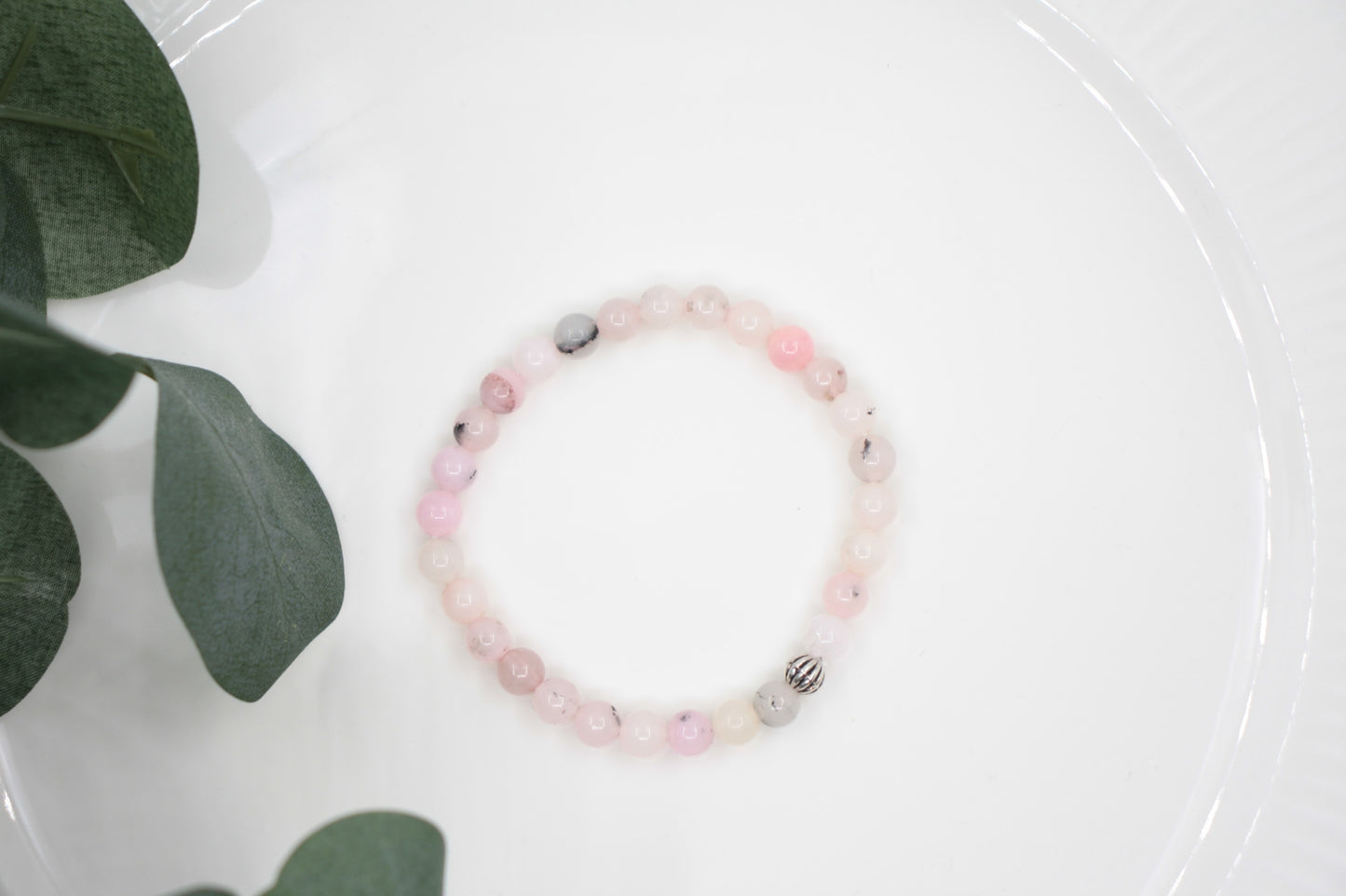 Jaspis kivihelmirannekoru vaaleanpunainen valkoisella alustalla. Vihreitä lehtiä taustalla. Pink jasper stone bead bracelet on a white surface. Green leaves in the background.