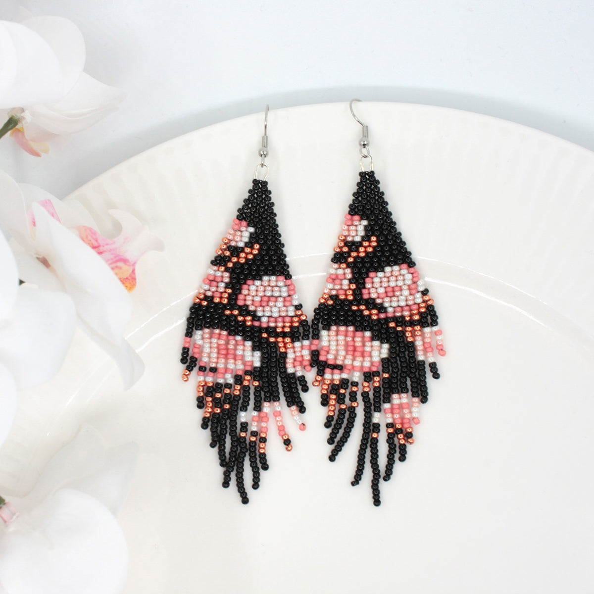 Long beaded seed bead fringe earrings on white plate. Base color of these earrings is and black and it has pink roses pattern.  Left side of the picture is white flowers. Pitkät käsintehdyt roikkuvat helmikorvakorut valkoisella lautasella. Pohjaväri näissä korvakoruissa on musta sekä, kuviona on pinkin sävyisiä ruusuja. Kuvan vasemmalla puolella on valkoisia kukkia.
